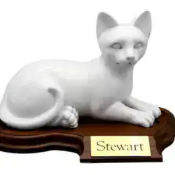 A white cat figurine lies on a dark wooden base with a gold nameplate that reads "Stewart." Perfect for honoring beloved pets, it’s an elegant tribute often chosen for pet cremation in Harris County, TX. The cat rests peacefully in a relaxed pose.