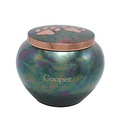 A metallic urn with a round shape and an iridescent finish, engraved "Cooper" on the front with paw prints on the lid, serves as a touching pet cremation memorial in Harris County, TX.
