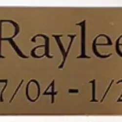 A bronze plaque reads "Raylee" with dates "7/04 - 1/21" engraved below in black letters, a loving tribute from pet cremation Harris County, TX.