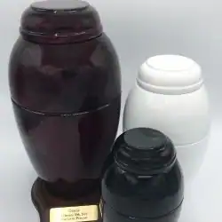 Three urns of varying sizes and colors—dark brown, white, and black—are displayed against a plain background. The largest urn, ideal for pet cremation in Harris County, TX, is mounted on a base featuring a small engraved plaque.