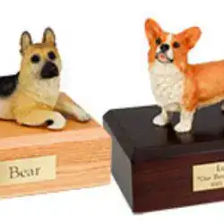 Two small dog figurines, one resembling a German Shepherd and the other a Corgi, are displayed on wooden bases with engraved nameplates—ideal as keepsakes for pet cremation in Harris County, TX.